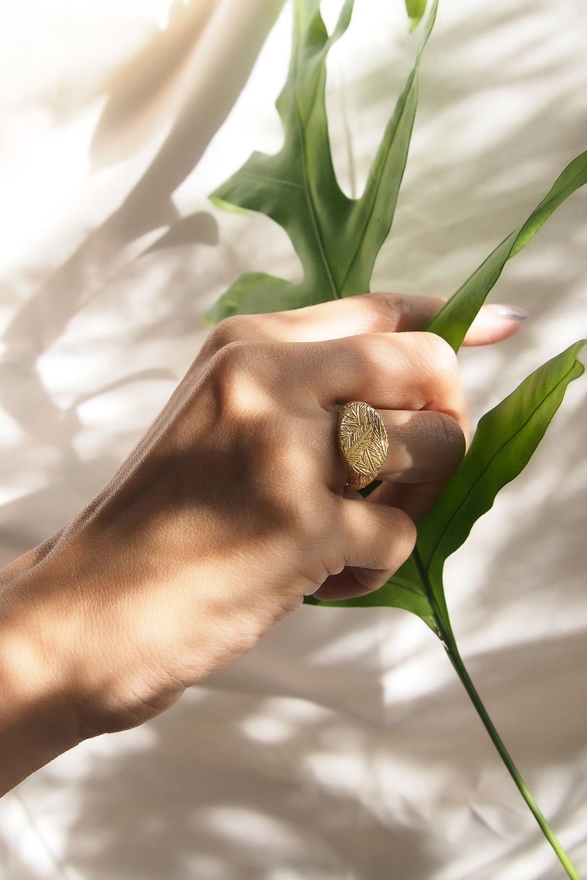 Close up of a hand wearing an elegant gold ring with distinct textures, featuring a feather pattern while holding a green leaf.