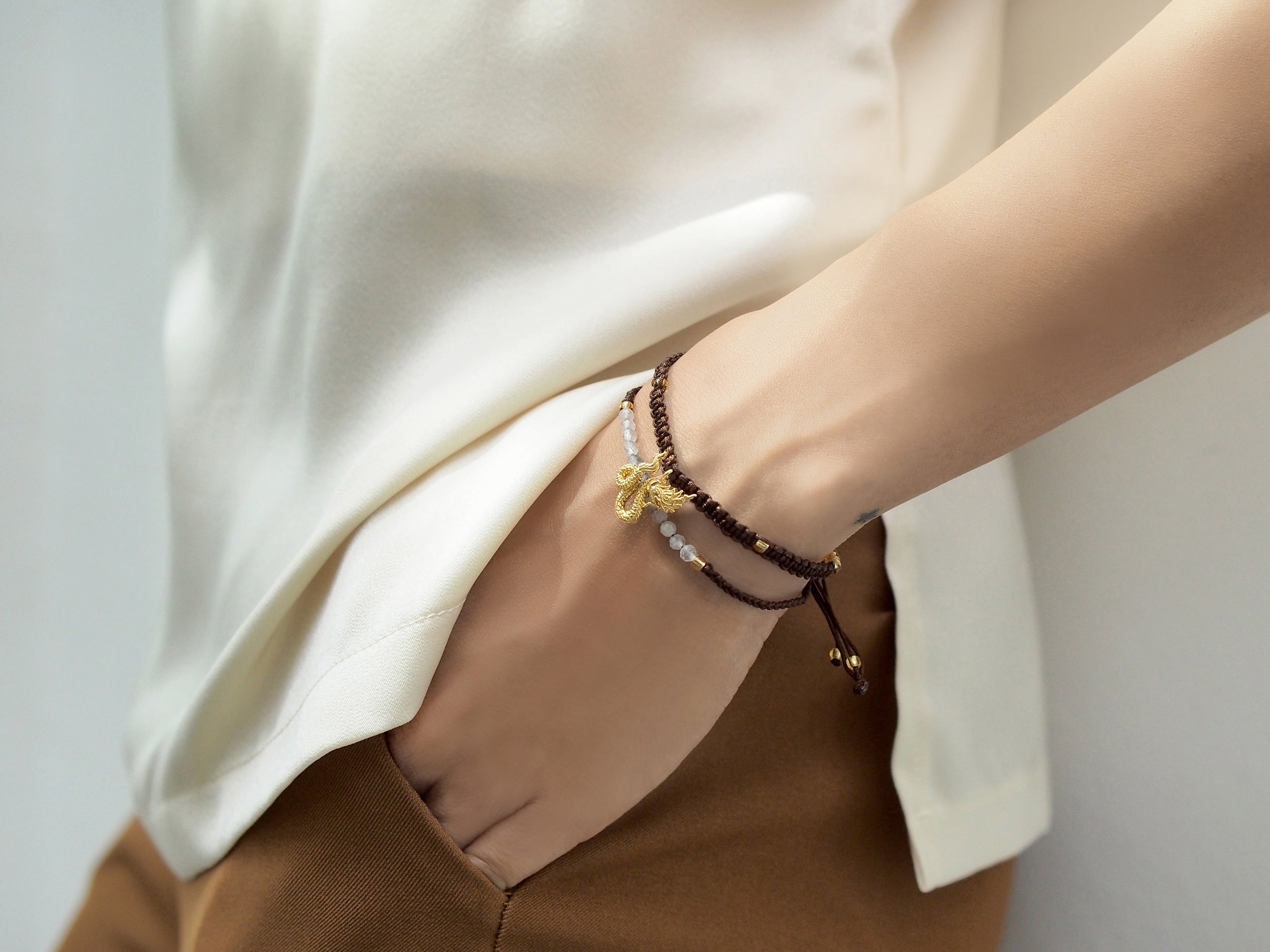 Close up of a hand adorned with set of two bracelets featuring a brown woven design with a gold Naga charm and a beaded grey and white bracelet.