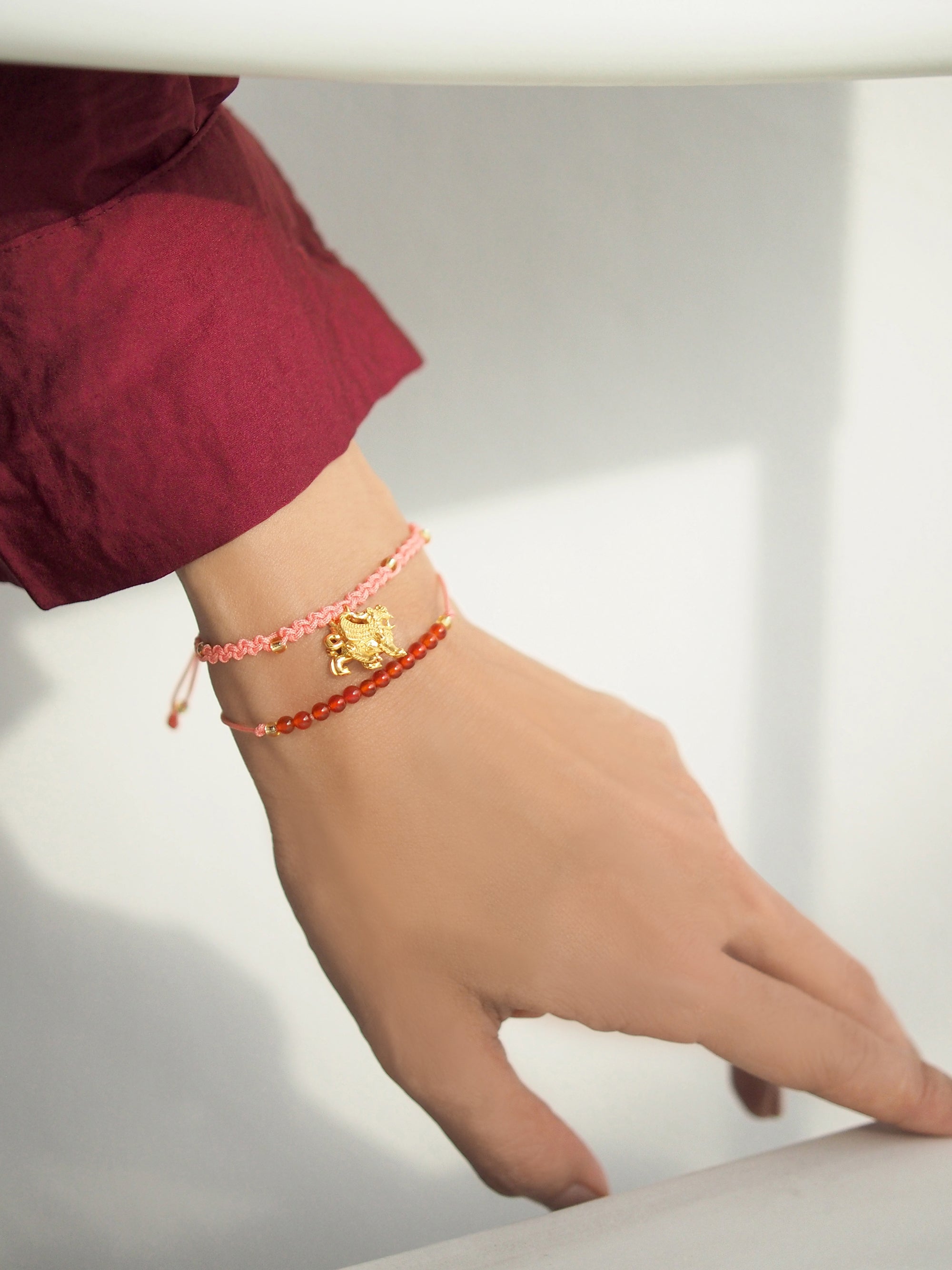 A close-up of a hand wearing a set of two pink bracelets: one with a gold Pixiu charm, the other with  red agate-round beads.