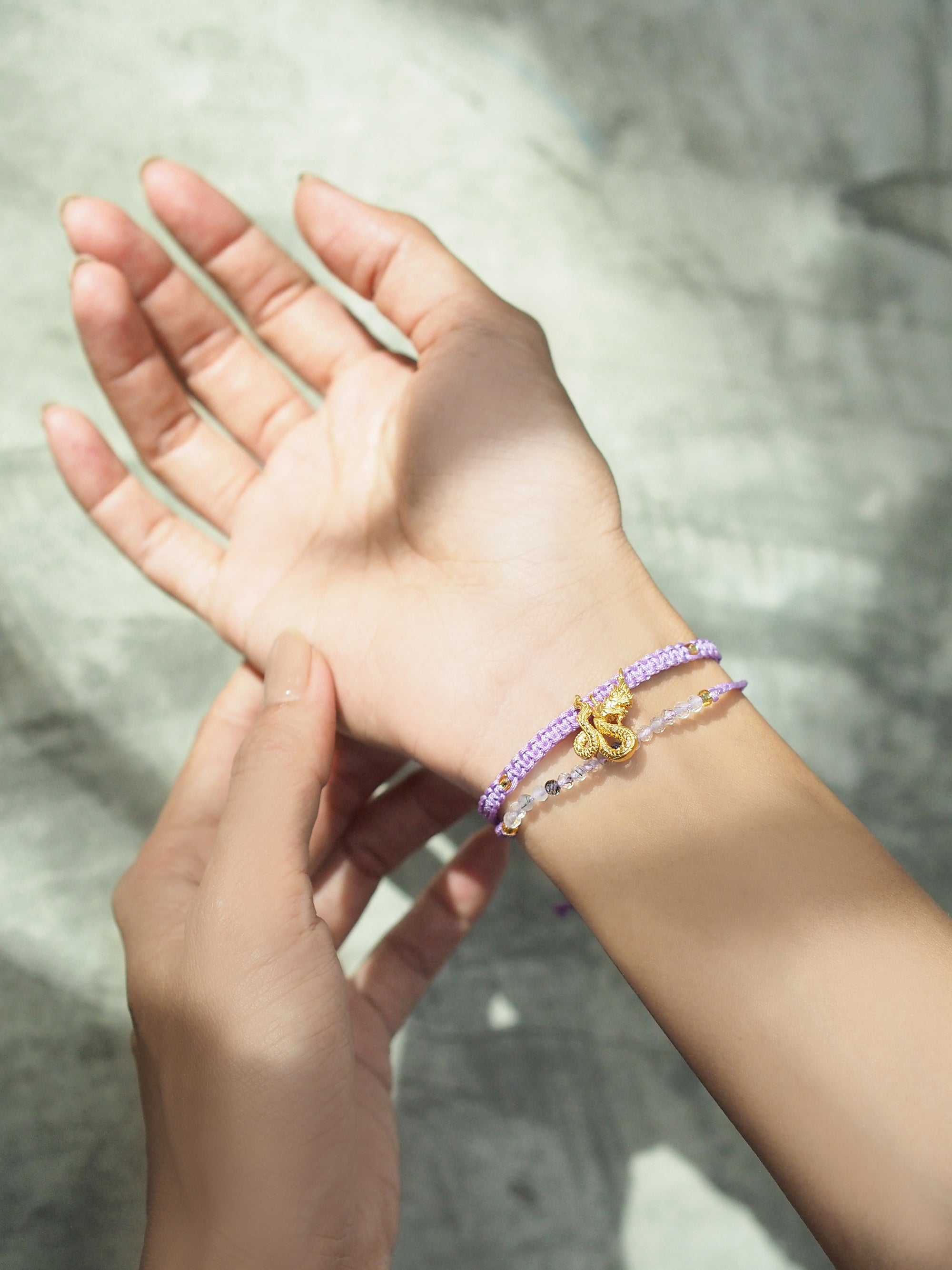 Close up of a hand adorned with set of two bracelets featuring a purple woven design with a gold Naga charm and a beaded grey and white bracelet.