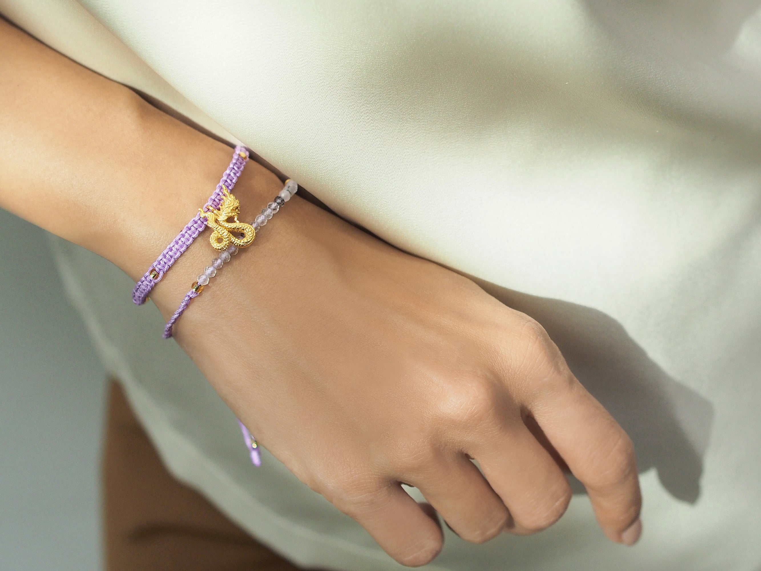 Close up of a hand adorned with set of two bracelets featuring a purple woven design with a gold Naga charm and a beaded grey and white bracelet.