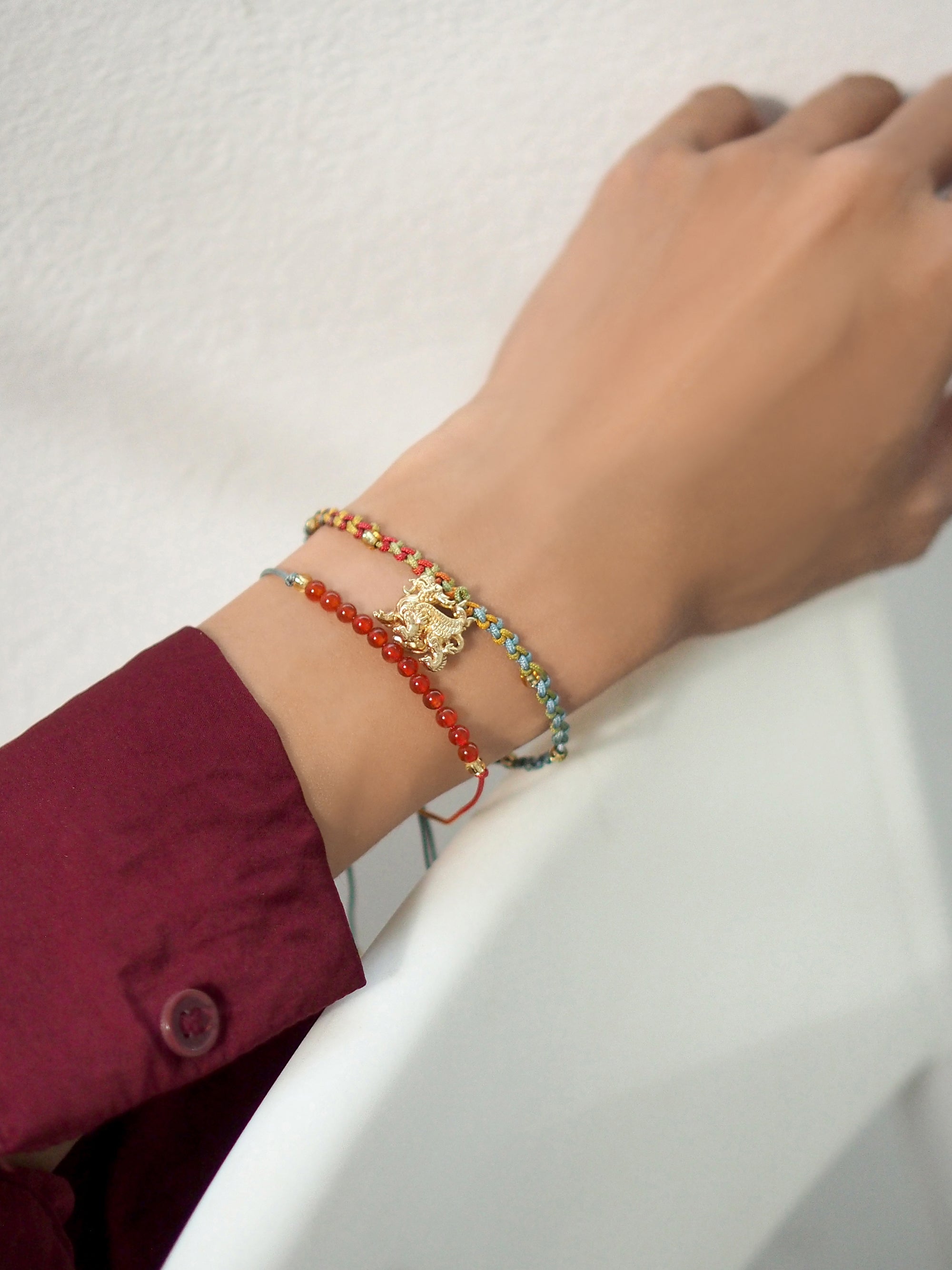 A close-up of a hand adorned with a set of two colorful bracelets: one with a gold Pixiu charm, the other with red agate round beads.