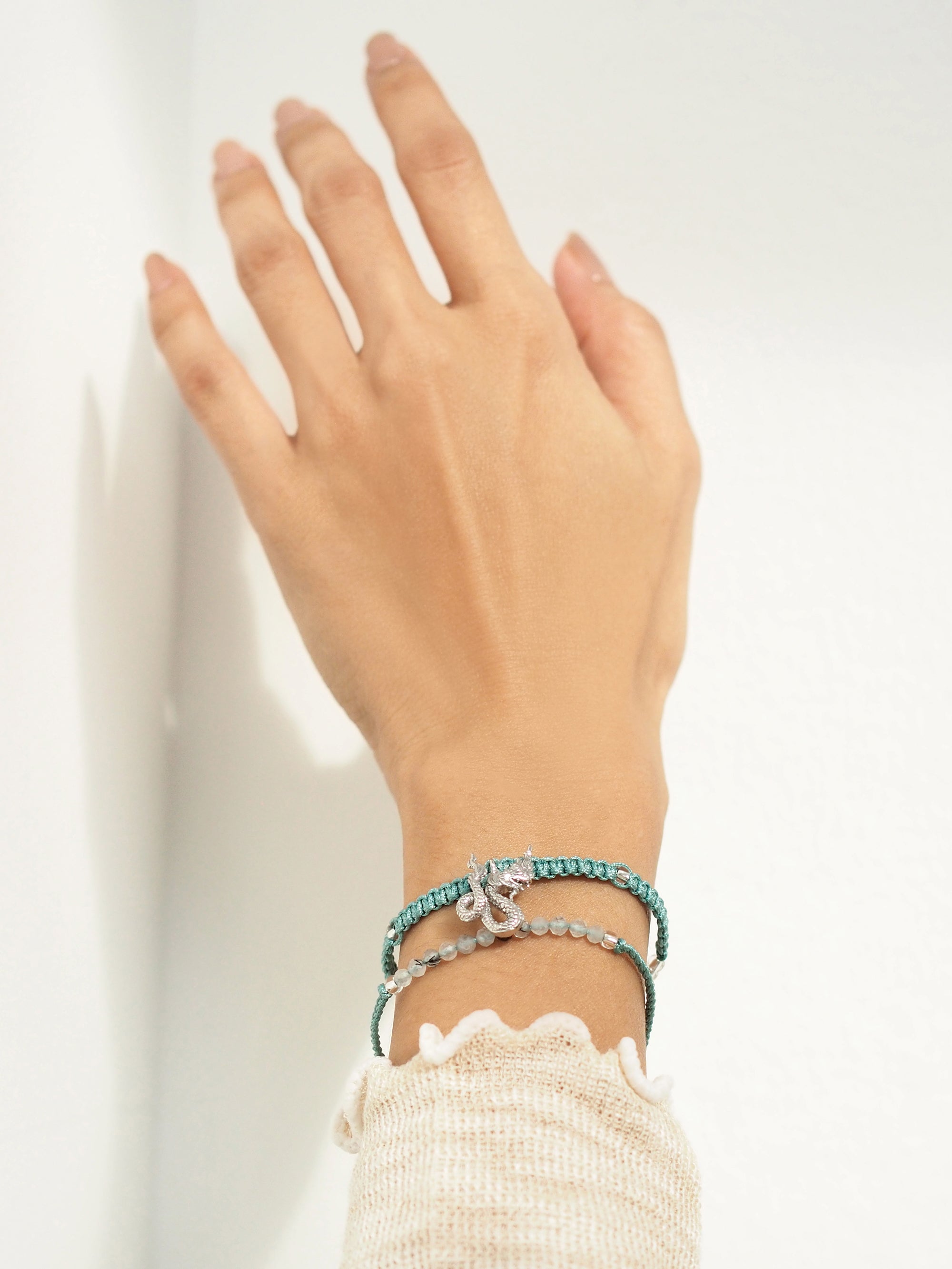 A close-up of a hand wearing a set of two light blue bracelets: one with a silver Naga charm, the other with grey quartz-faced beads.