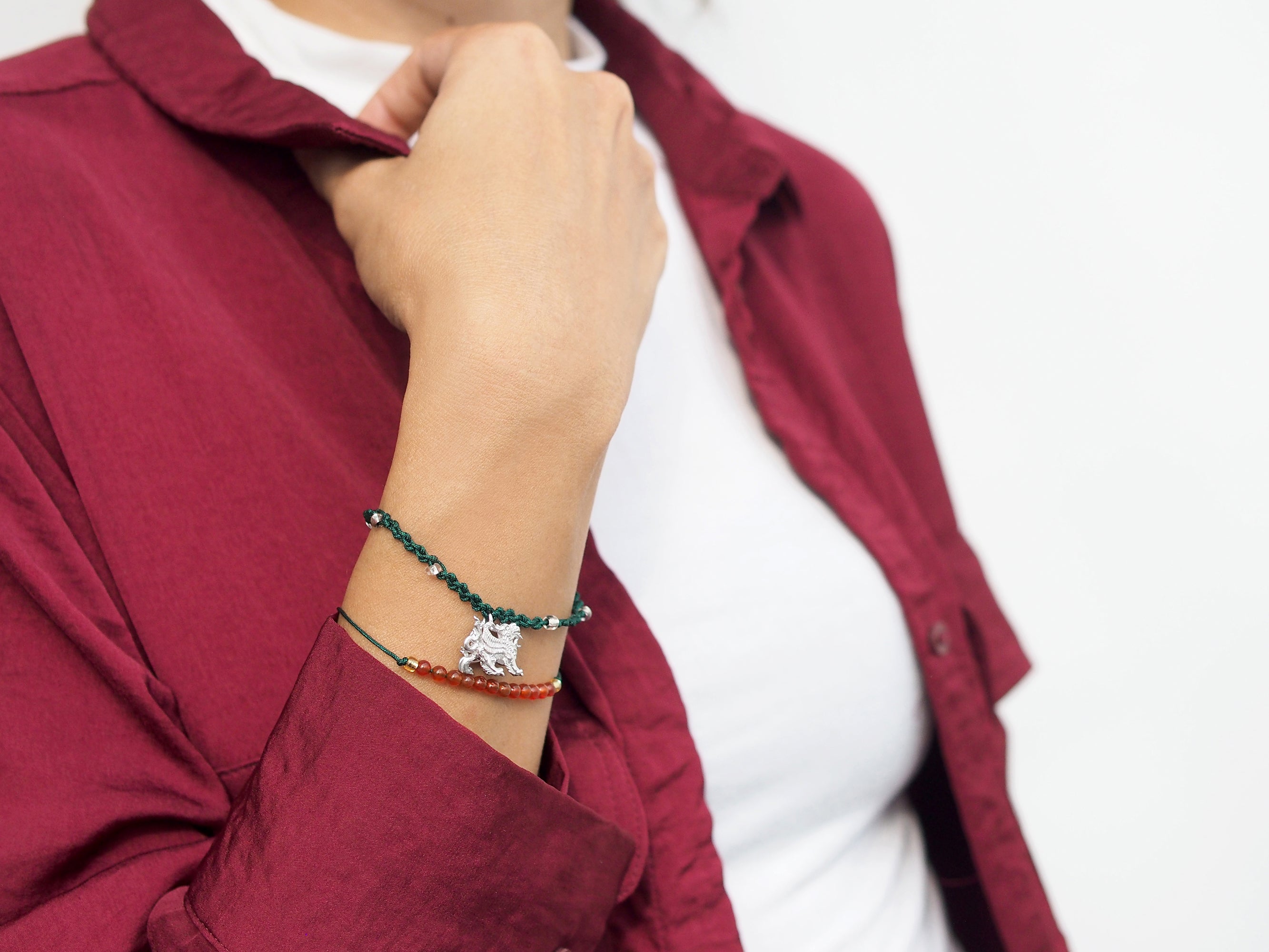 A close-up of a hand wearing a set of two green bracelets: one with a silver Pixiu charm, the other with  red agate-round beads.