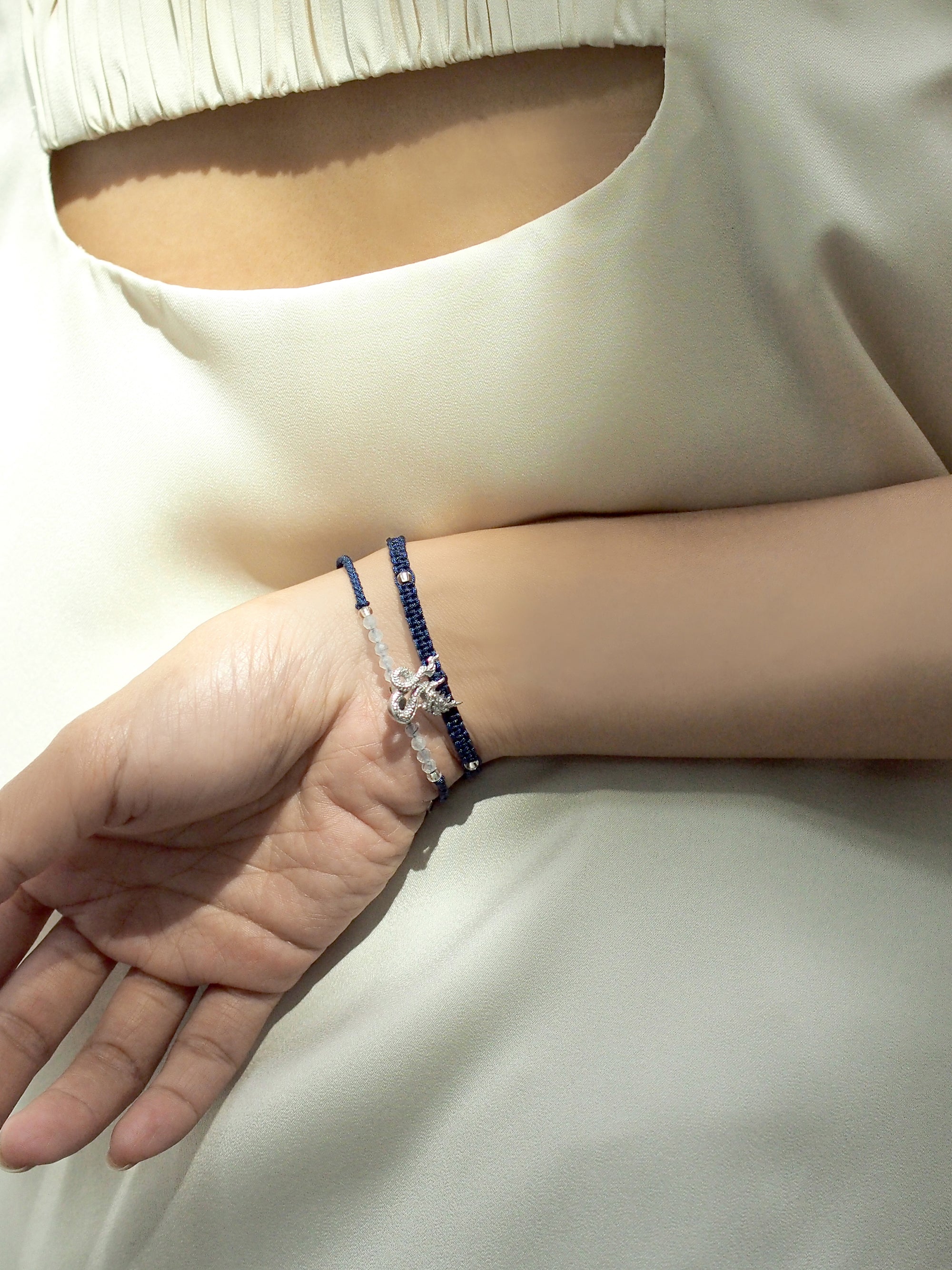 A close up of a hand wearing a set of two navy blue bracelets: one with a silver Naga charm, the other with grey quartz-faced beads.