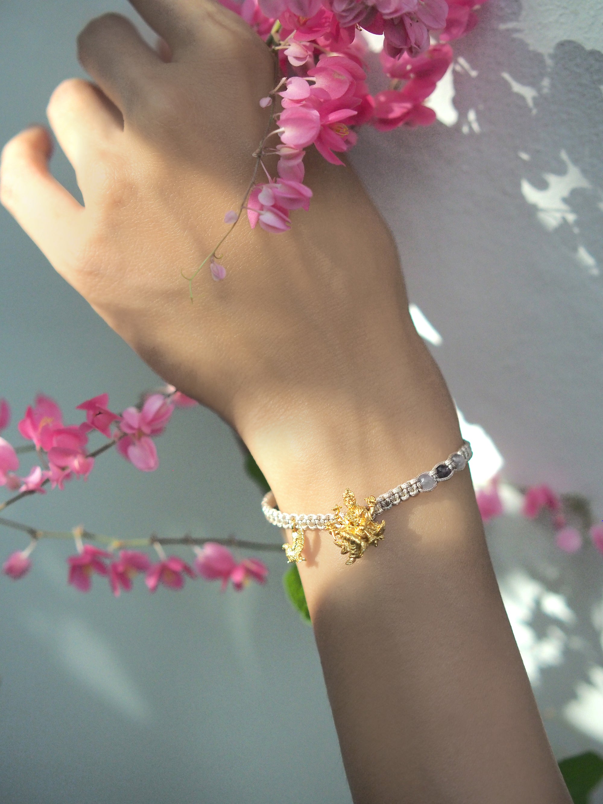 A hand with a bracelet with Trimurti Vishnu and Garuda charms holding pink flowers.