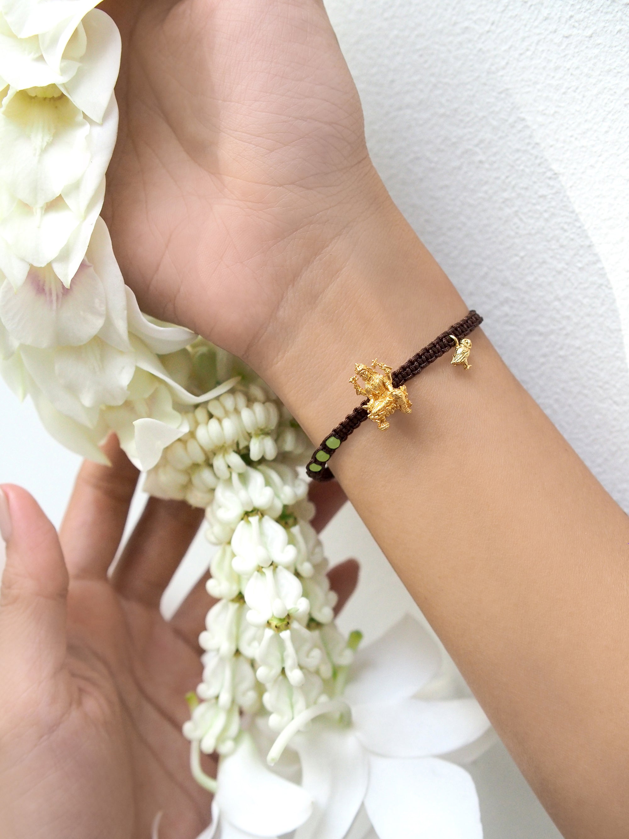 Close-up of a hand wearing a charm bracelet featuring Tridevi Lakshmi and owl charms, while holding a garland.