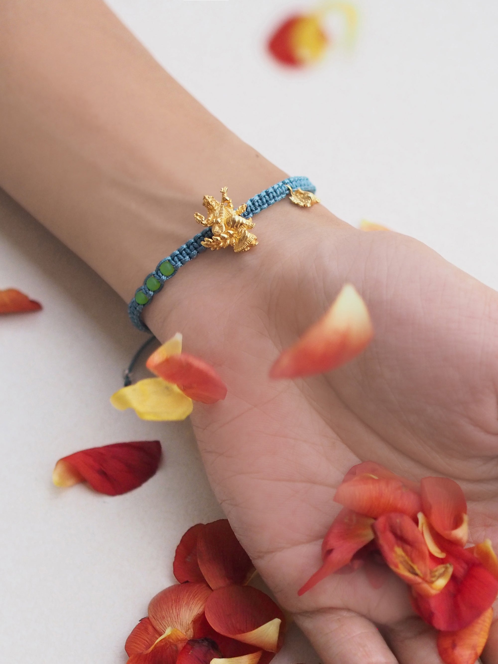 Close-up of a green bracelet adorned with a gold Tridevi Sarasvati charm worn on wrist amidst scattered flower petals.