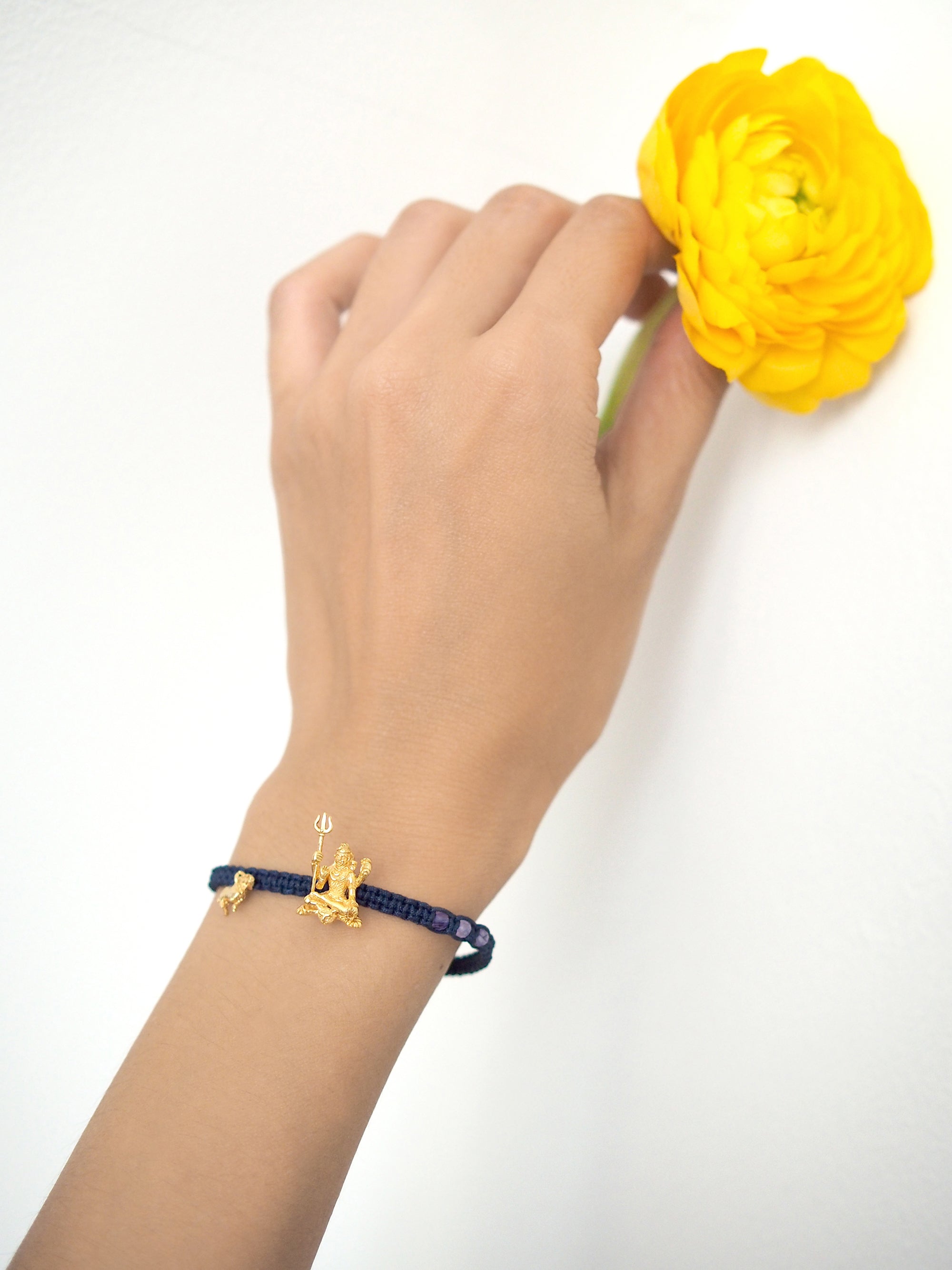 A hand adorned with a Trimurti Shiva charm bracelet with amethyst-faced beads holding a yellow flower.