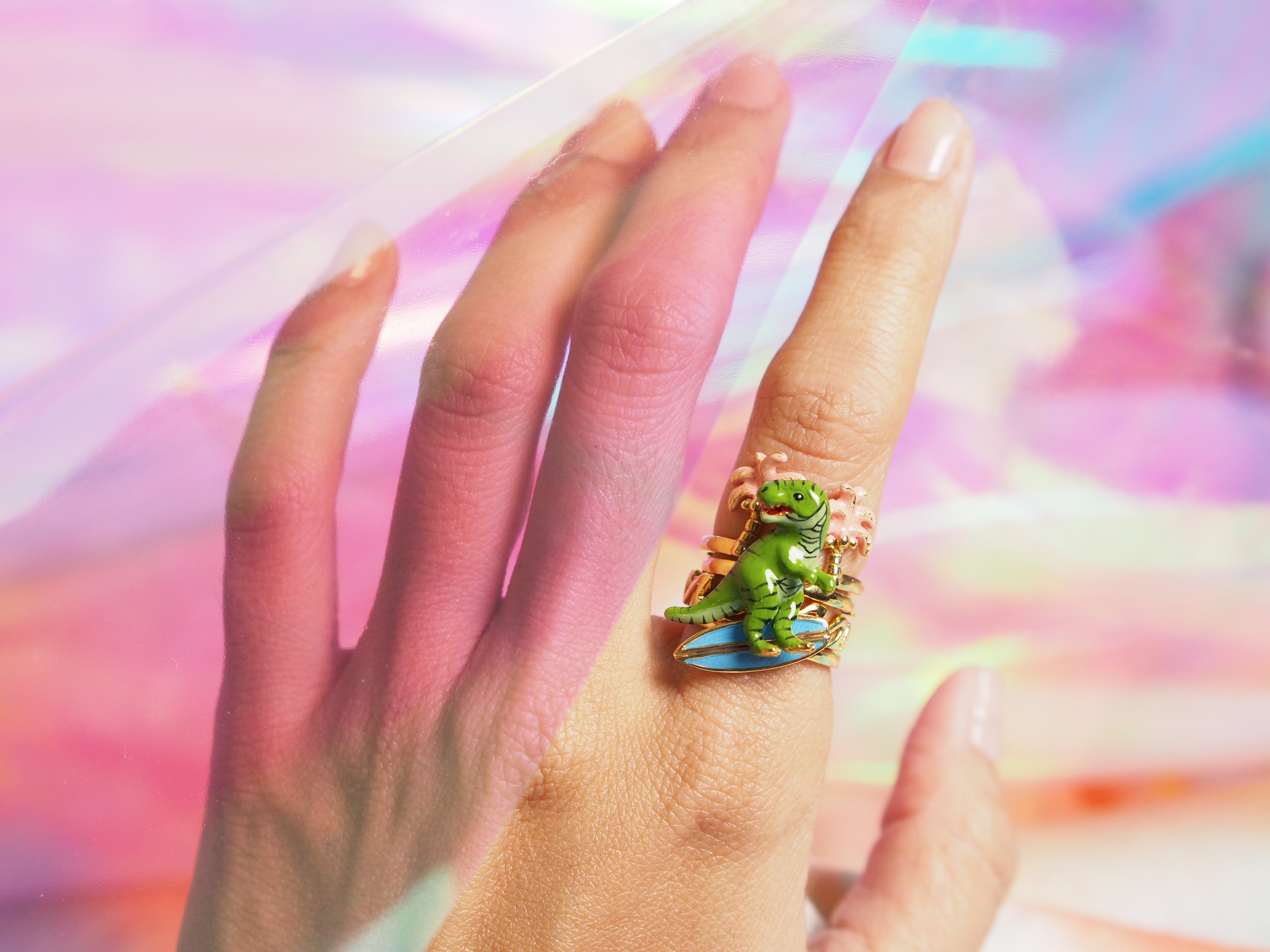 Close-up of a charming stacking ring fearing a green dinosaur, palm trees, a surfboard, and a wave-pattern design worn on a hand against a colorful background.