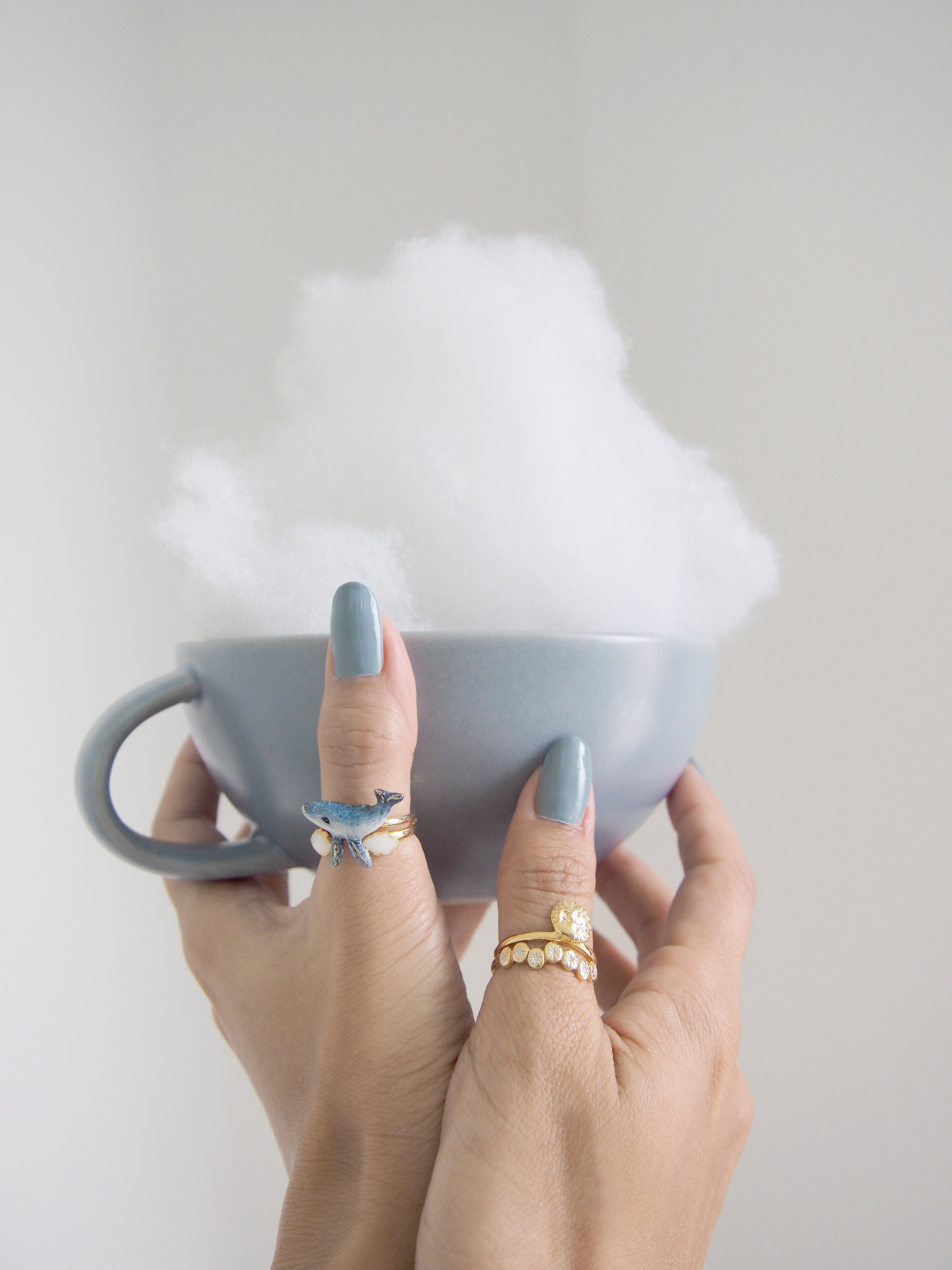 A cup filled with fluffy white cotton resembling clouds, held by hands adorned with whimsical rings.