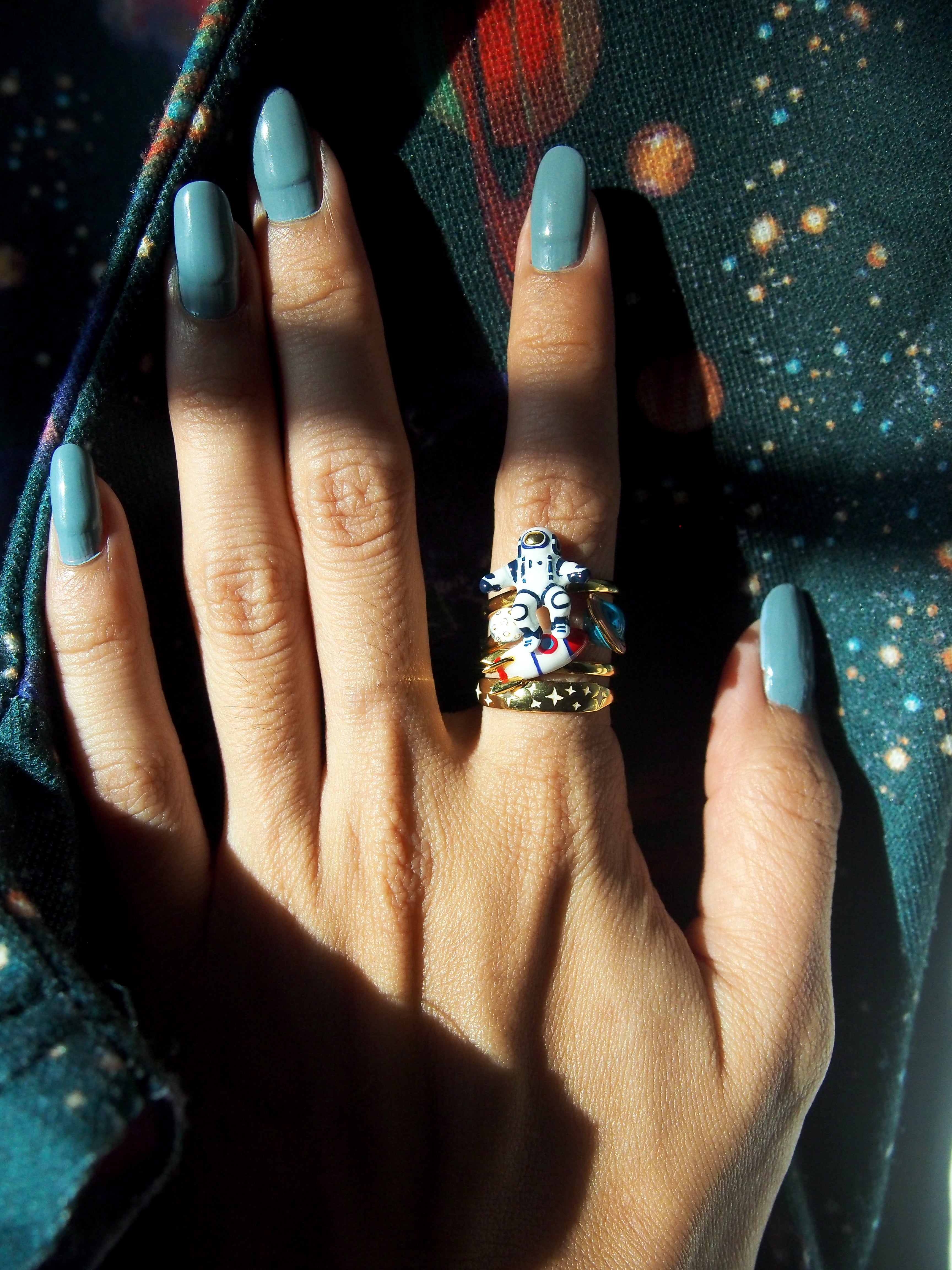 Space-inspired stacking ring on a hand with painted nails, surrounded by a colorful cosmic-patterned background.