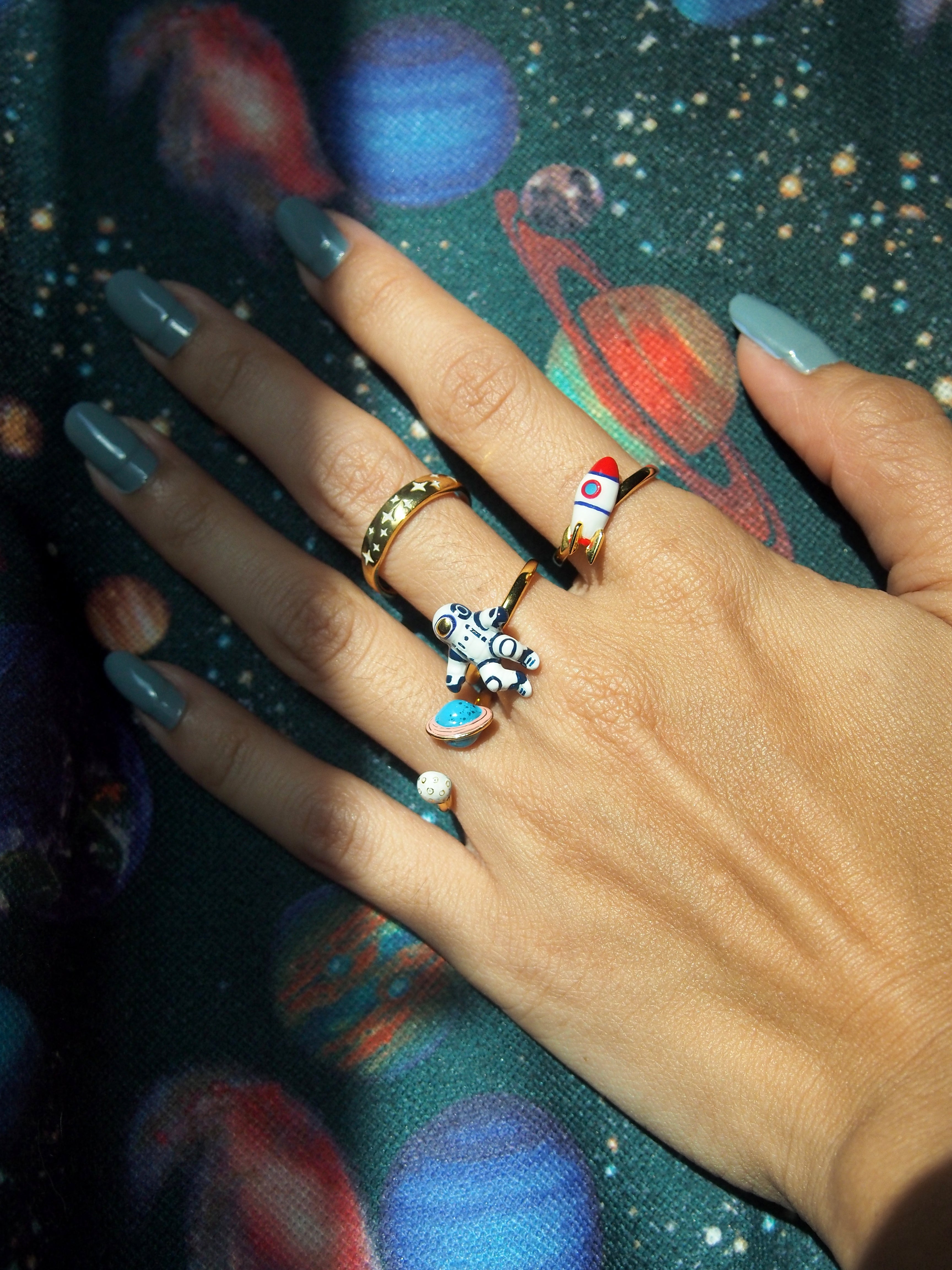 Four space-themed rings on a hand with painted nails, surrounded by a colorful cosmic-patterned background.