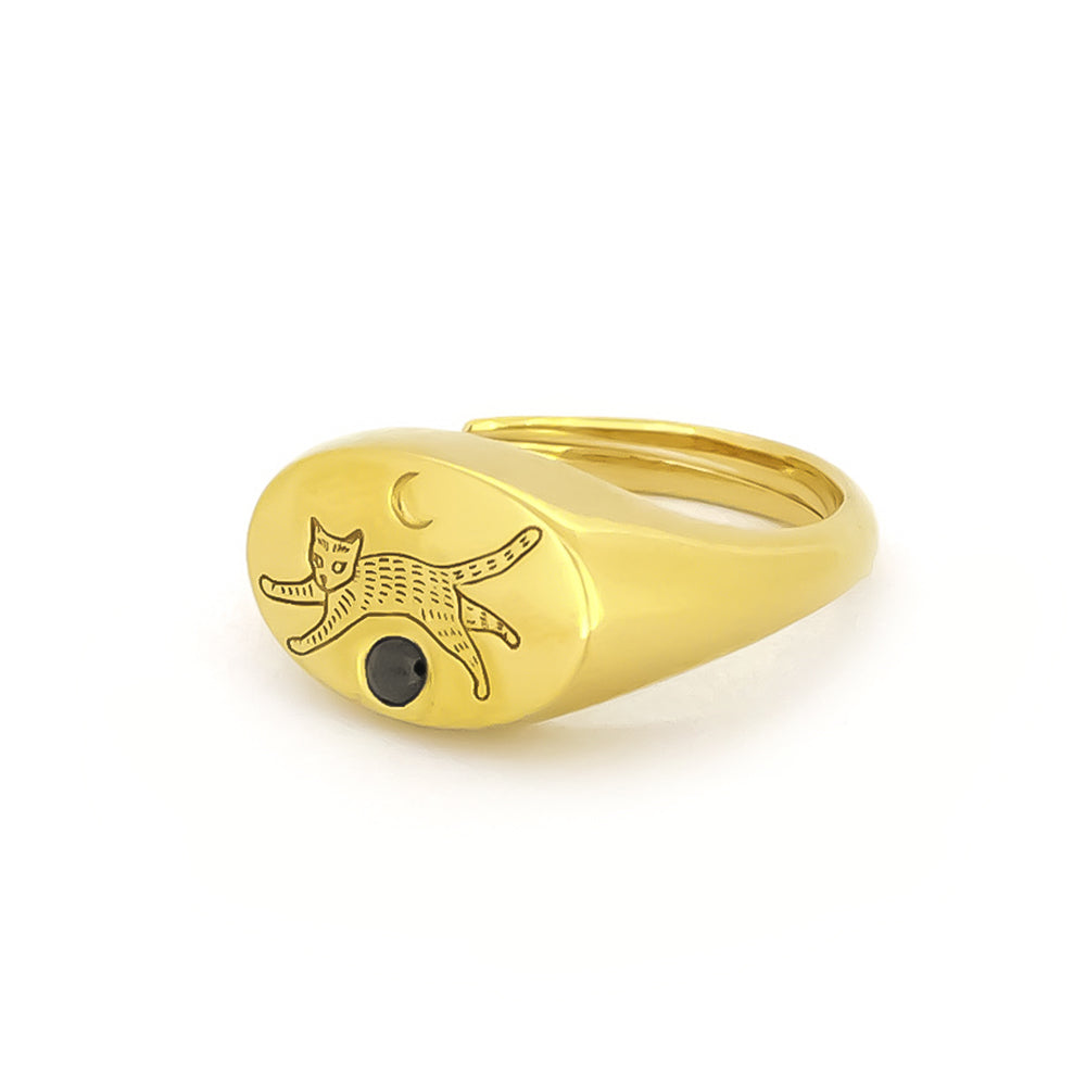 Gold oval-shaped ring featuring an engraved playful cat design and a crescent moon, complemented by a small black gemstone.