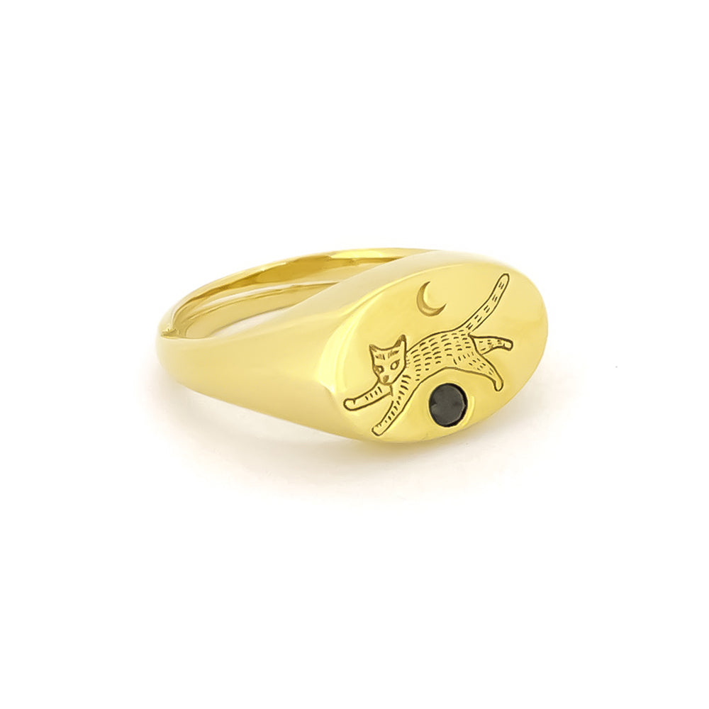 Gold oval-shaped ring featuring an engraved playful cat design and a crescent moon, complemented by a small black gemstone.