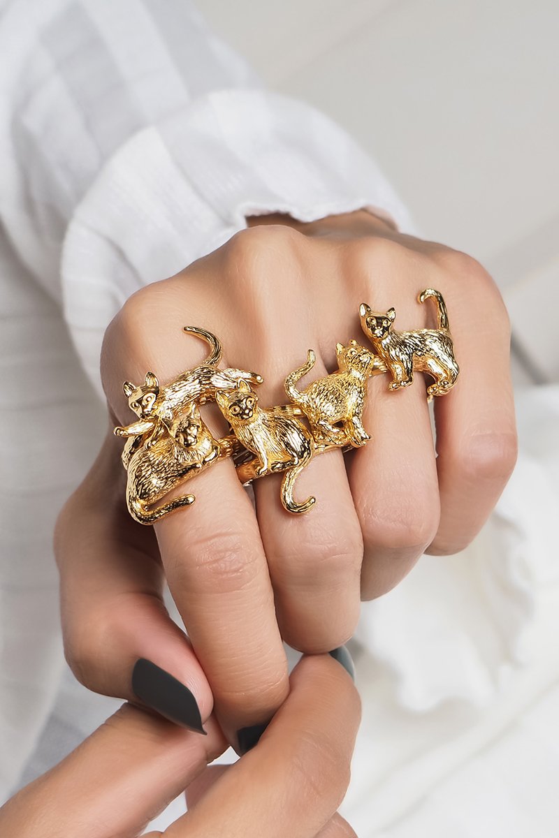 A hand adorned with multiple gold rings featuring cat designs in various poses, resting on the light-colored setting background.