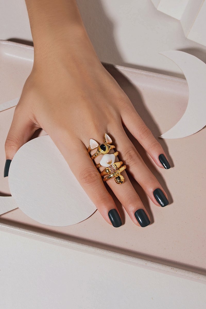 Whimsical rings with cat features, including ears, eyes, nose, paws and a bell, displayed on a hand with dark nail polish.