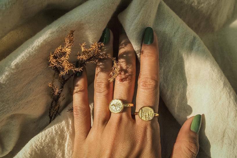 Close up of a hand wearing two gold rings featuring an engraved forest scene and mountain, resting on a fabric.