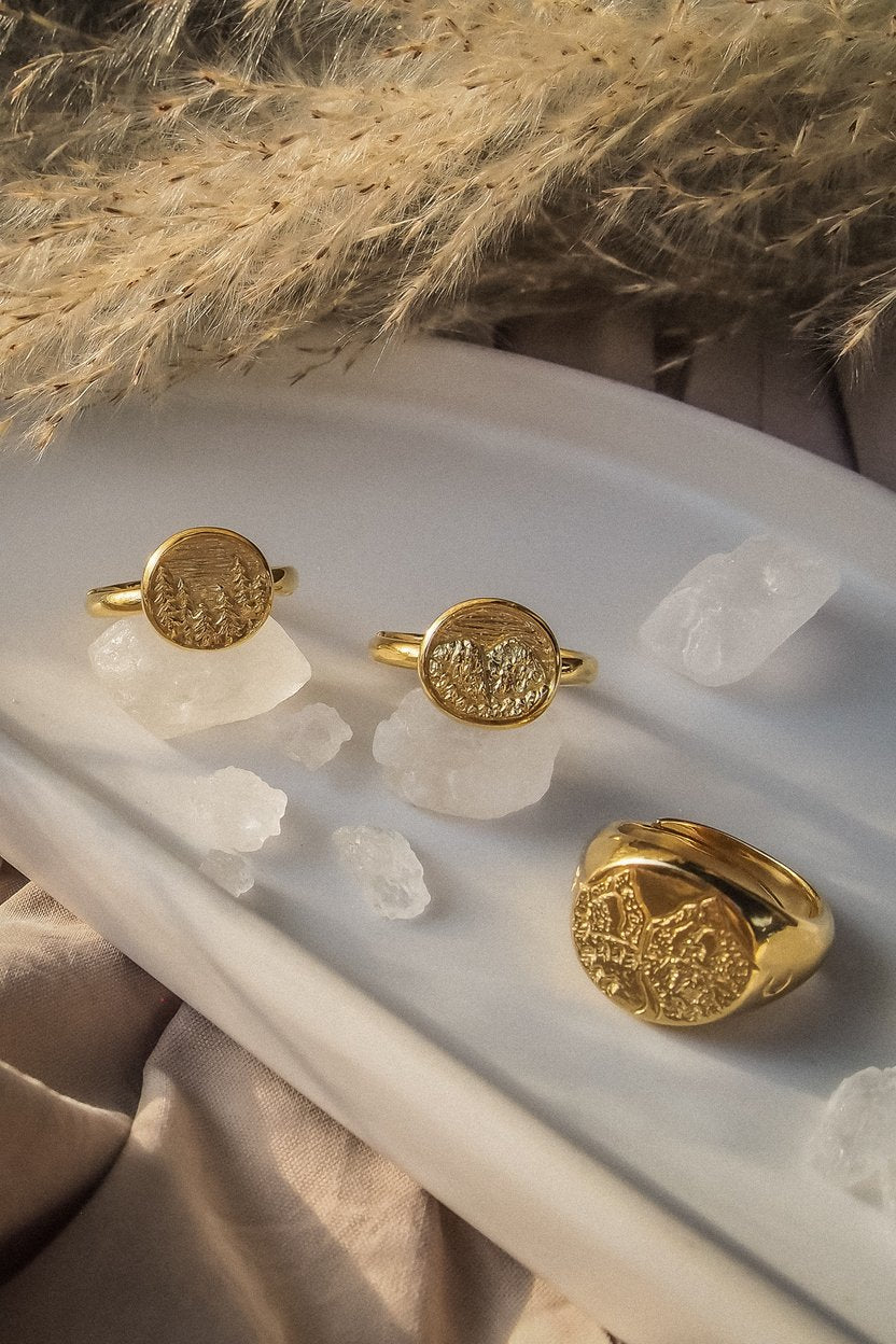 Nature-inspired three gold rings featuring landscape of forest and mountain placed on a plate with icy decorative stones and dried flowers.